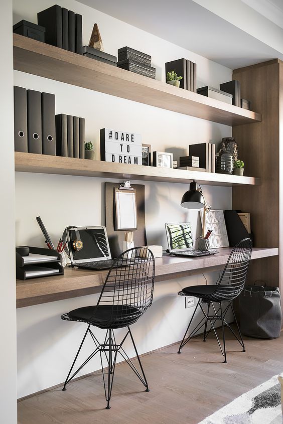 a large niche with built in shelves and a double desk, some books, potted plants, a black table lamp and black chairs