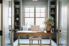 a light grey modern farmhouse home office with built-in shelves, a stained trestle desk, a neutral chair, a printed rug and a chandelier