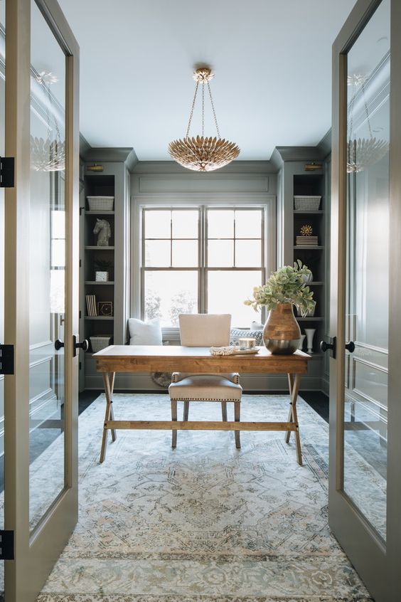 a light grey modern farmhouse home office with built-in shelves, a stained trestle desk, a neutral chair, a printed rug and a chandelier