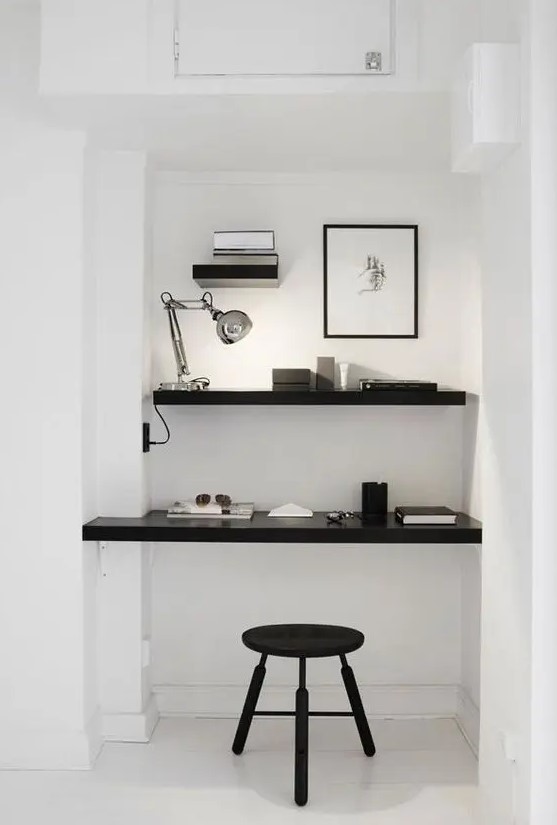 a minimalist niche with a built in shelf and desk, a table lamp, some laconic decor and a black stool