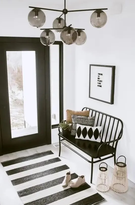 a modern black and white foyer with a black glass door, a black bench and a striped rug, an artwork and lanterns