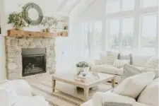 a modern country living room designed in neutrals, with a fireplace clad with stone, cozy furniture and printed textiles