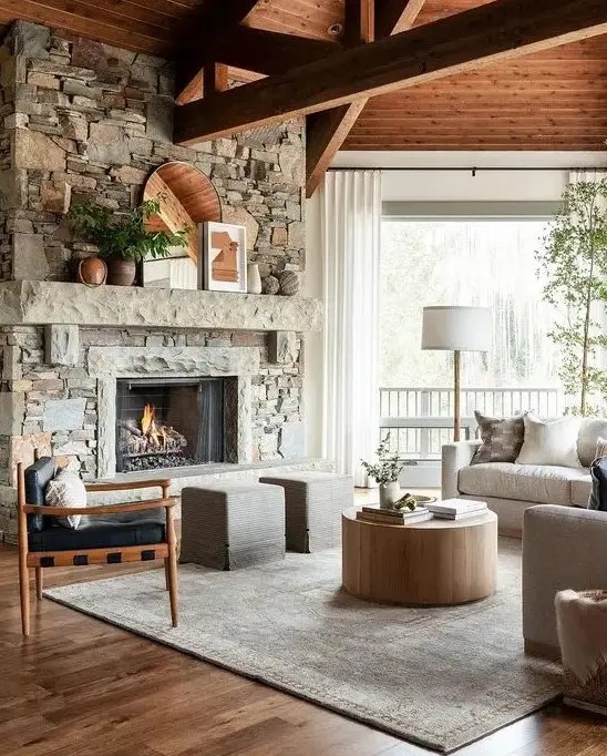 a modern country living room with a fireplace clad with faux stone, wooden beams, neutral furniture and a round coffee table