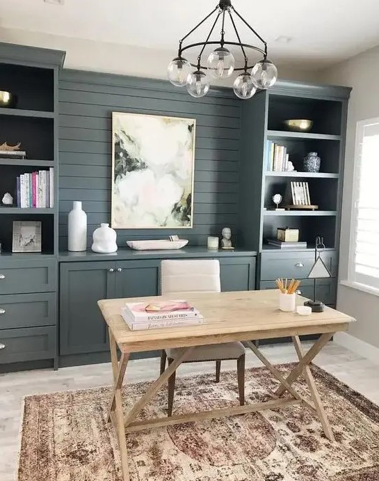 a modern country working space with slate grey storage units that are built in, a wooden trestle desk and a chandelier plus a boho rug