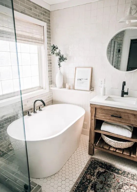a modern farmhouse bathroom with grey and white tiles, a stained vanity, an oval tub, a shower space, a round mirror and some decor