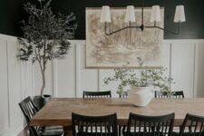a modern farmhouse black and white dining room with black walls and creamy paneling, a stained table, black chairs, a chandelier, greenery