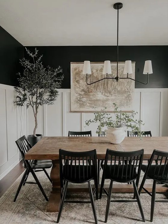 a modern farmhouse black and white dining room with black walls and creamy paneling, a stained table, black chairs, a chandelier, greenery