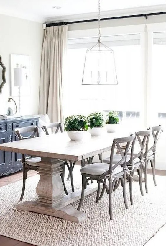 a modern farmhouse dining space with a black credenza, a heavy dining table, stained chairs, a pendant lamp and some potted greenery