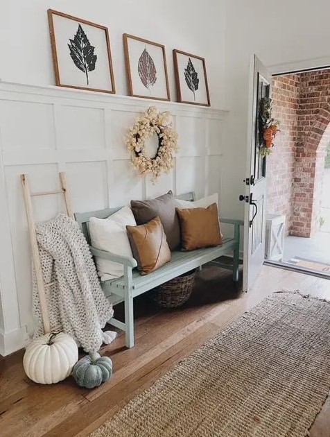 a modern farmhouse entryway with a white paneled wall, a mint bench with pillows and a blanket, a gallery wall and some pumpkins