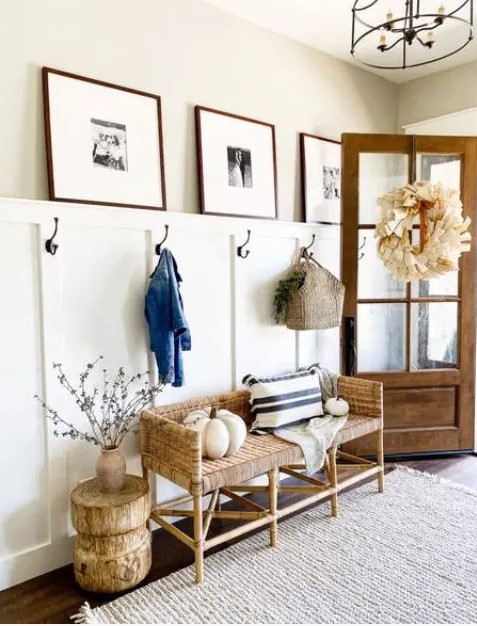 a modern farmhouse entryway with a woven bench, a paneled wall, a side table of wood, a gallery wall and some pumpkins