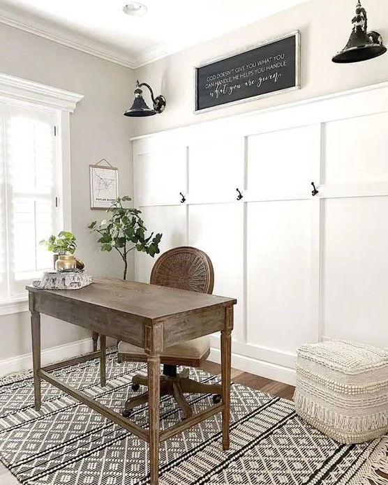 a modern farmhouse home office with a paneled wall, a stained desk and a chair, a boho pouf, black vintage sconces and greenery