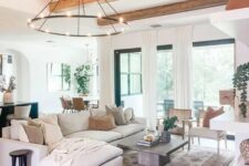a modern farmhouse living room with wooden beams, a chandelier, a neutral sectional, a coffee table, a fireplace, some chairs