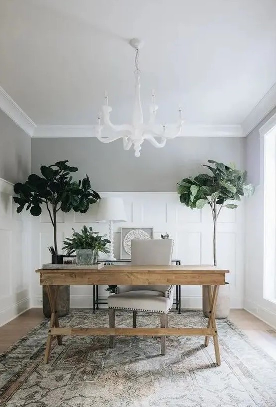 a modern home office with a white chandelier, a wooden trestle desk, a white chair, a printed rug and statement plants
