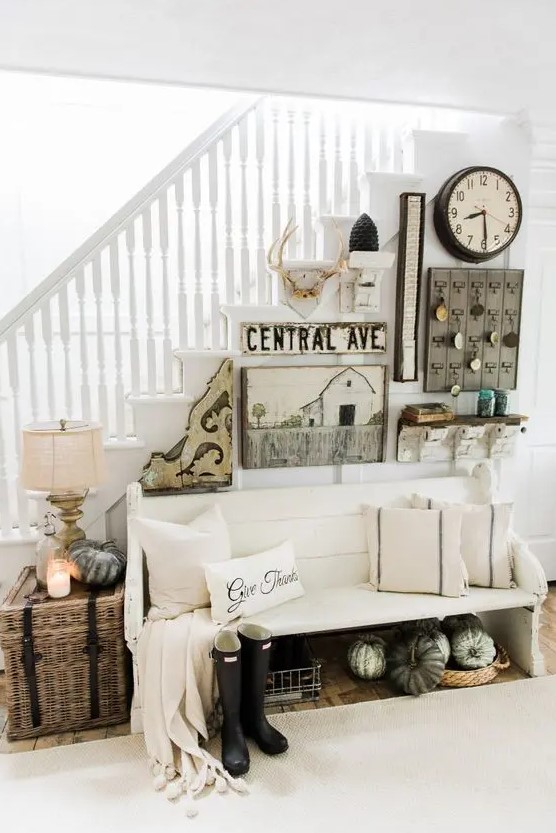 a modern neutral farmhouse entryway with a white bench, a basket chest with a lamp, a gallery wall in rustic style and pumpkins