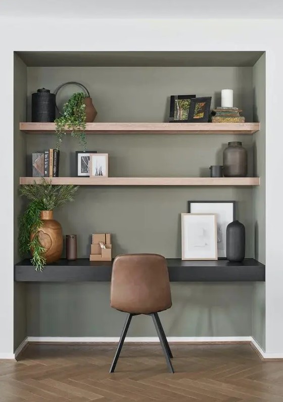 a modern niche painted olive green, with built-in shelves and a black desk, some decor and greenery, a brown leather chair and artwork