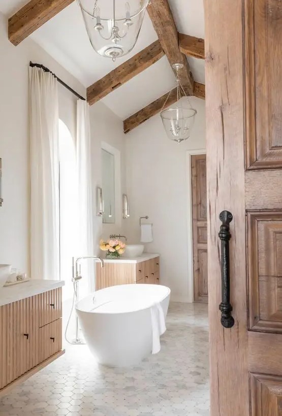 a neutral modern farmhouse bathroom with fluted floating vanities, an oval tub, a tiled floor and stained wooden beams