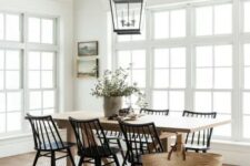 a neutral modern farmhouse dining room with a stained table and black chairs, a black frame pendant lamp, a woven rug and some art and greenery
