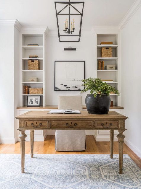 a neutral modern farmhouse home office with built in shelves, a stained vintage desk, a neutral chair, a potted plant and a pendant lamp