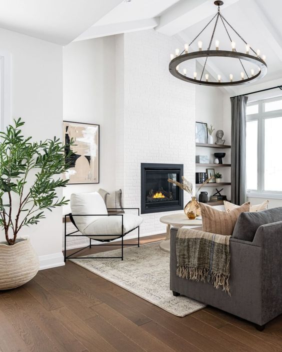 a neutral modern farmhouse living room with a built-in fireplace, dark-stained shelves, a taupe sofa, white chairs and a round coffee table