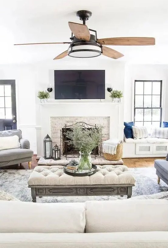 a neutral modern farmhouse living room with a fireplace clad with brick, neutral furniture and built-in daybeds, an upholstered ottoman and a fan
