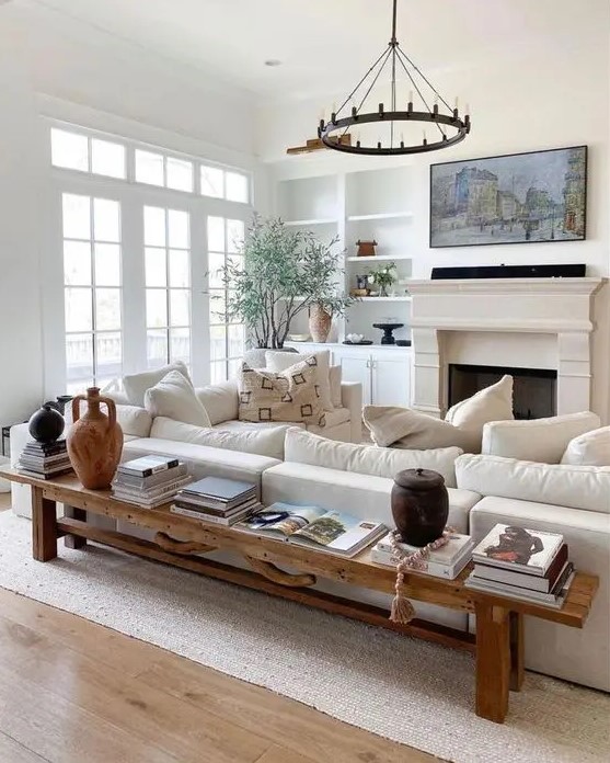 a neutral modern farmhouse living room with built-in shelves, a fireplace, a creamy sofa, a bench as a coffee table and a chandelier