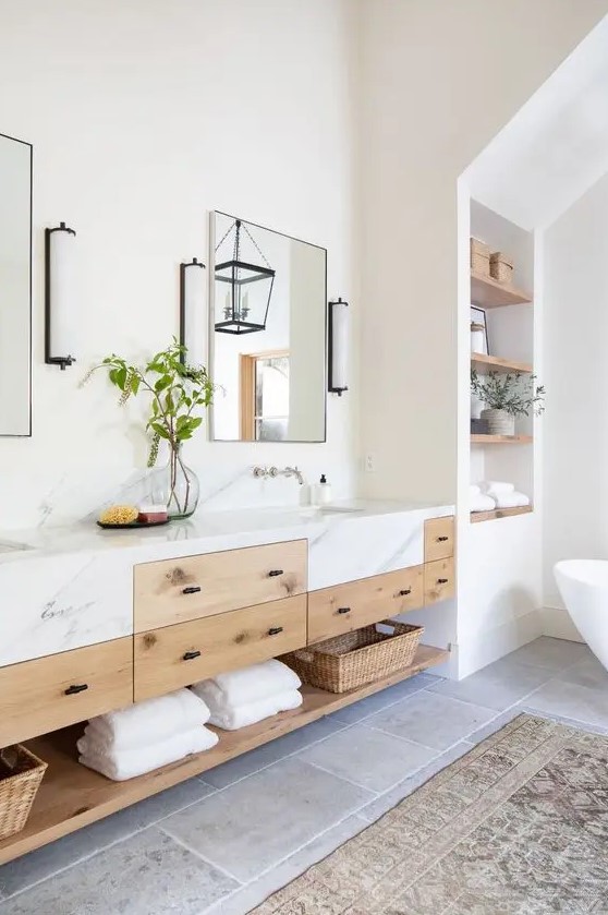 a refined modern farmhouse bathroom with an oval tub, built in shelves, a double timber vanity with a marble countertop, mirrors and sconces
