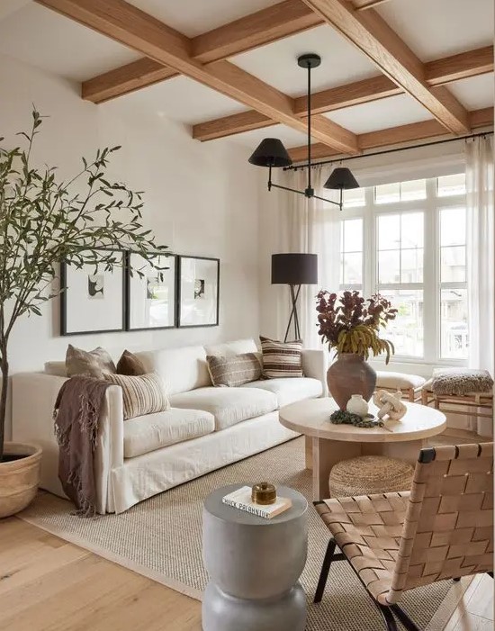 a serene modern farmhouse living room with a coffered ceiling, a creamy sofa and pillows, stools, a woven chair, a coffee table and a black chandelier
