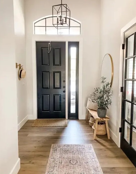 a simple black and white entryway with a black door, a stained bench, a round mirror, a printed rug and a potted plant