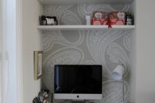 a small niche home office with printed wallpaper, built-in shelves and a desk, some necessary stuff and a clear chair