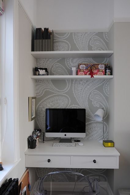 a small niche home office with printed wallpaper, built in shelves and a desk, some necessary stuff and a clear chair