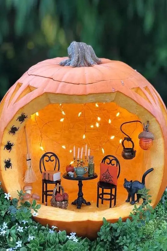 a stylish Halloween pumpkin diorama with lights, small black chairs and a table with candles, a kettle, a black cat and some black spiders