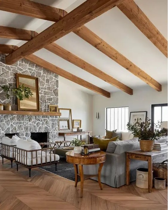 a welcoming modern farmhouse living room with wooden beams, a fireplace clad with stone, a grey sofa and white chairs, stained and refined furniture