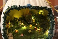 a white Halloween diorama with grass, pebbles, lights, flowers and a tiny house inside plus some hay