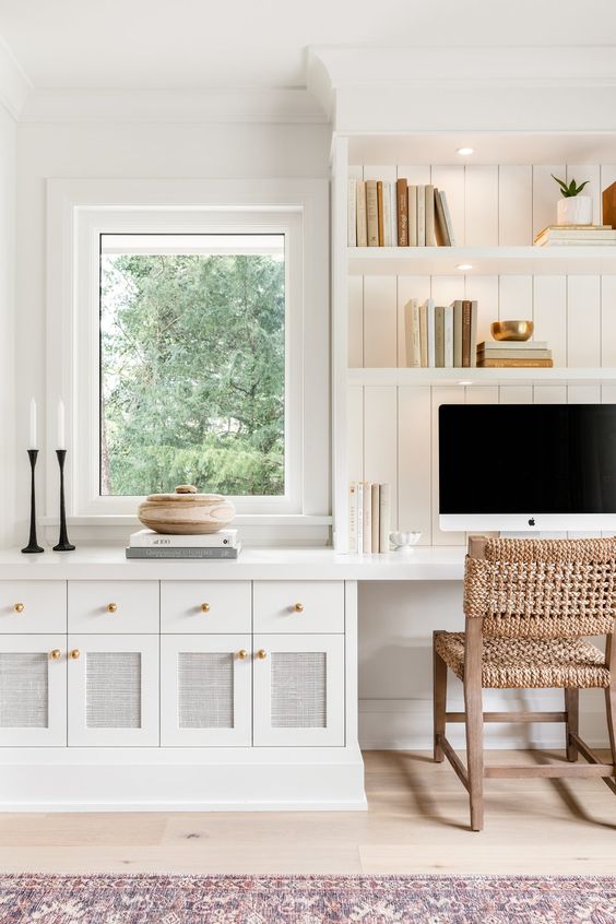 a white modern farmhouse home office with shiplap walls, built-in cabinets and shelves, a woven chair and some decor