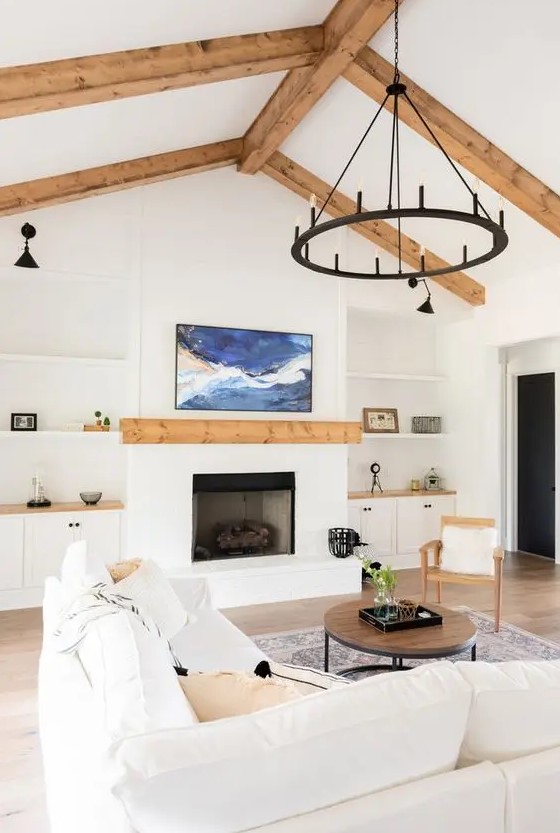 a white modern farmhouse living room with wooden beams, a fireplace, built-in shelves and cabinets, a creamy sofa and a black chandelier