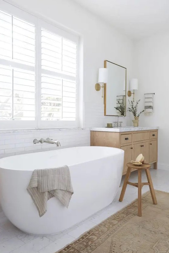 an airy farmhouse bathroom with an oval tub, a timber vanity and a wooden stool, a window with shutters and a boho rug