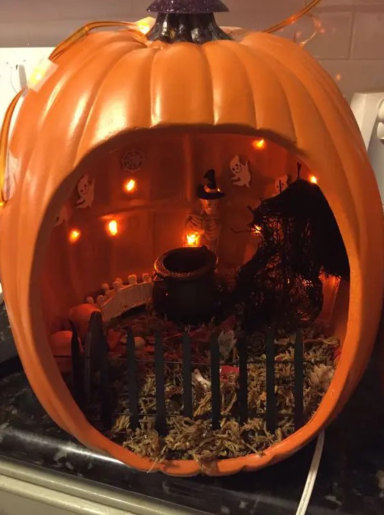 an orange pumpkin diorama with hay, a fence, lights, a cauldron, a skeleton in a witch hat and some lights