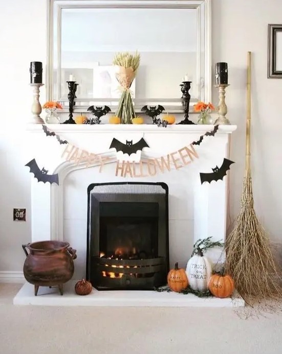 a Halloween mantel with bats, candles, pumpkins, orange blooms, a wheat bundle and some pumpkins and a broom on the floor