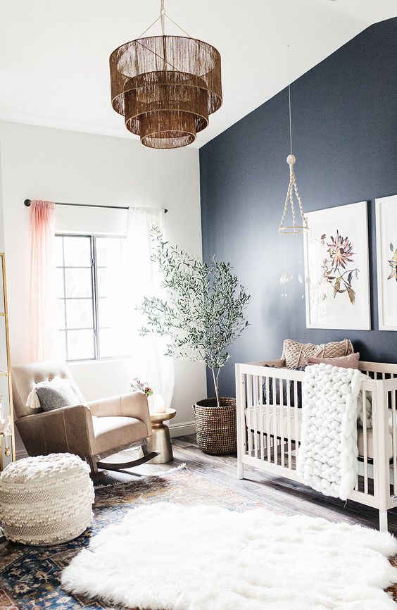 a boho nursery with a navy accent wall, a white crib, layered rugs, a rocker chair, pouf, a potted plant and a gallery wall