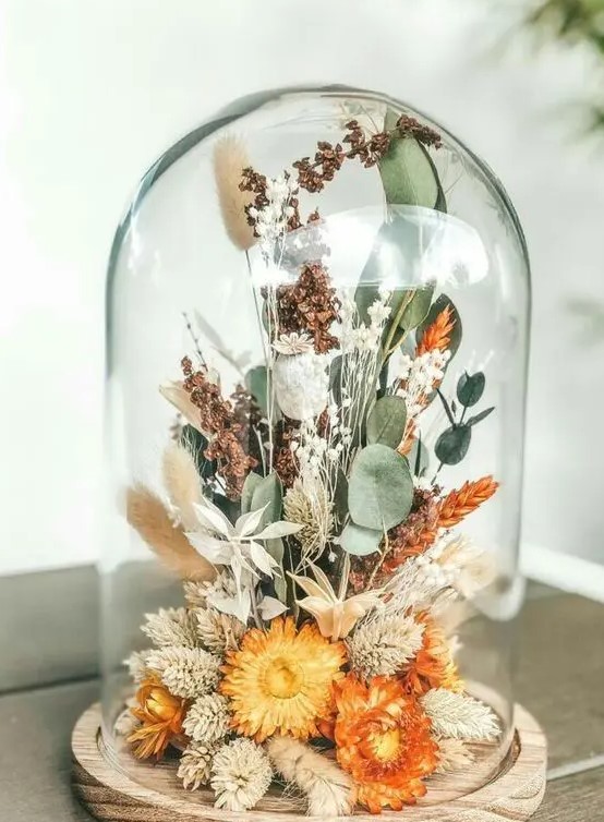 a cloche with a wooden base, with orange dried blooms, grasses, greenery feels like summer and can be a nice decoration for the fall, too