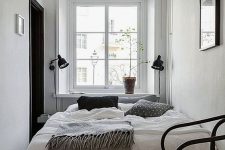 a narrow Scandinavian bedroom with a bed, black sconces and a black chair, some greenery and an artwork