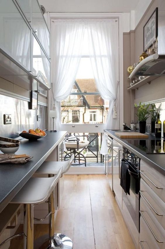 a narrow kitchen with neutral cabinets, black countertops, tall stools, ledges and shelves plus a view through the balcony entrance