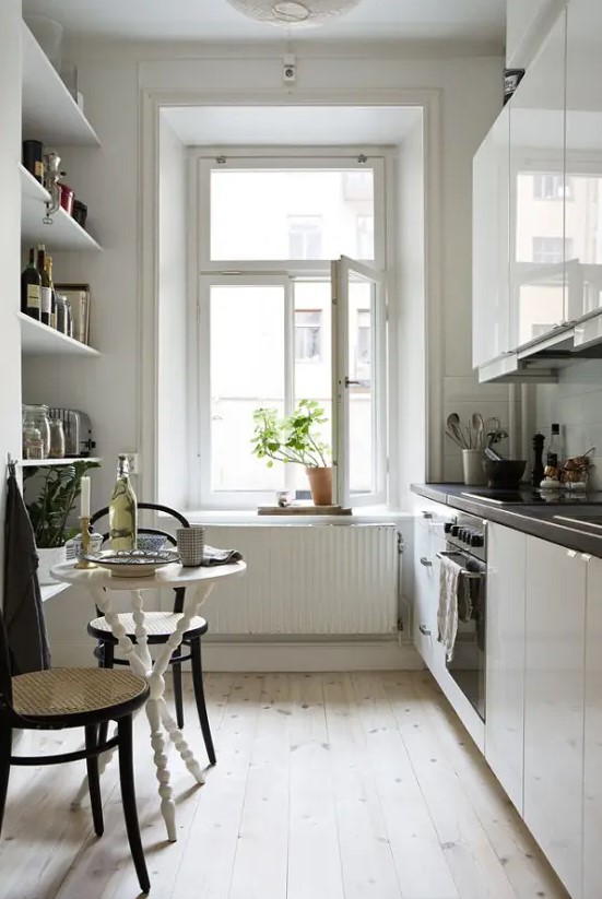 a narrow kitchen with open shelves, sleek white cabinets, black countertops, a small table and cane chairs plus potted greenery