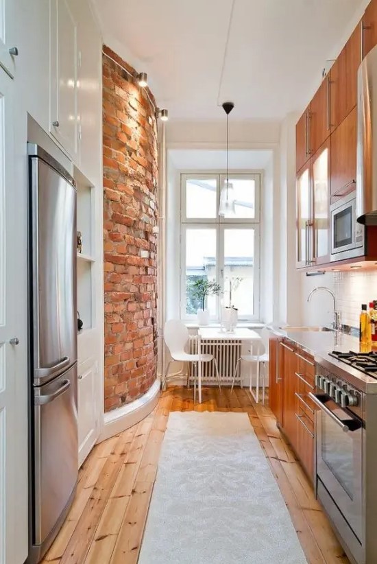 a narrow kitchen with rich stained cabinets, built in appliances, a brick wall, a white dining set and a stained floor