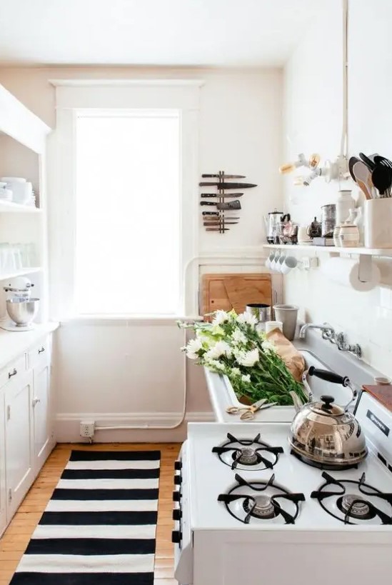 a narrow kitchen with white cabinets, a sink and a cooker, a printed rug, some appliances and greenery
