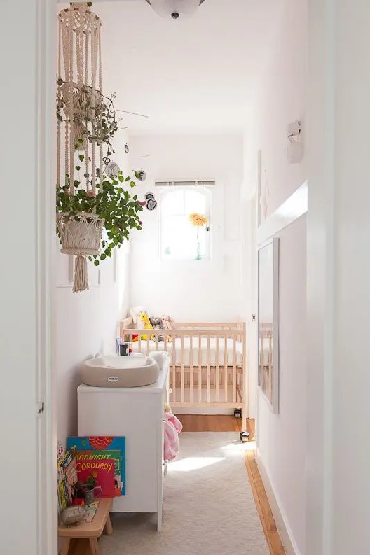 a narrow nursery in white, with a stained crib, a white dresser, some windows and artwork, greenery and toys
