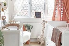 a neutral boho nursery with a large white crib, a changing table, a chair, bookshelves, a coral canopy and some lovely decor