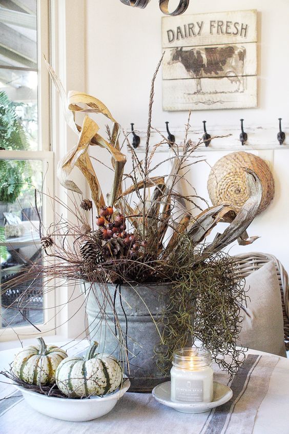 a relaxed rustic fall decoration of a bucket with twigs, pinecones, dried husks and pumpkins and candles around is cozy and cool
