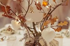 a rustic fall centerpiece of a glass vase with pinecones, branches with leaves and twigs, ornaments, bells and clay ornaments