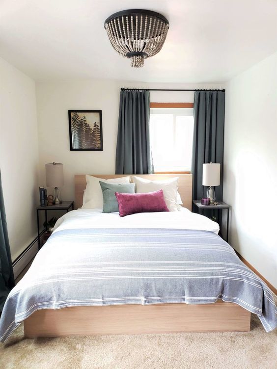 A small and narrow bedroom with a light stained bed and neutral bedding, nightstands with lamps and a beaded chandelier